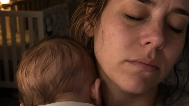 A mother at night with her eyes closed, her baby's head resting against her cheek, a crib behind them.