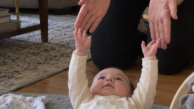 A baby on a play mat on the living-room floor, both arms reaching up toward his mother's open hands.