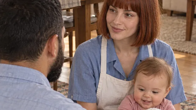 A mother on a living-room rug smiling at the father, their baby in her lap.