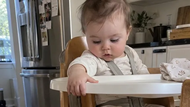 A baby in a wooden high chair in a sunny kitchen, looking down past the tray at the toy she just let go of.