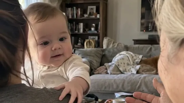 A baby on her mother's shoulder in the living room, one hand holding on, her grandmother's open hands reaching in from the right.